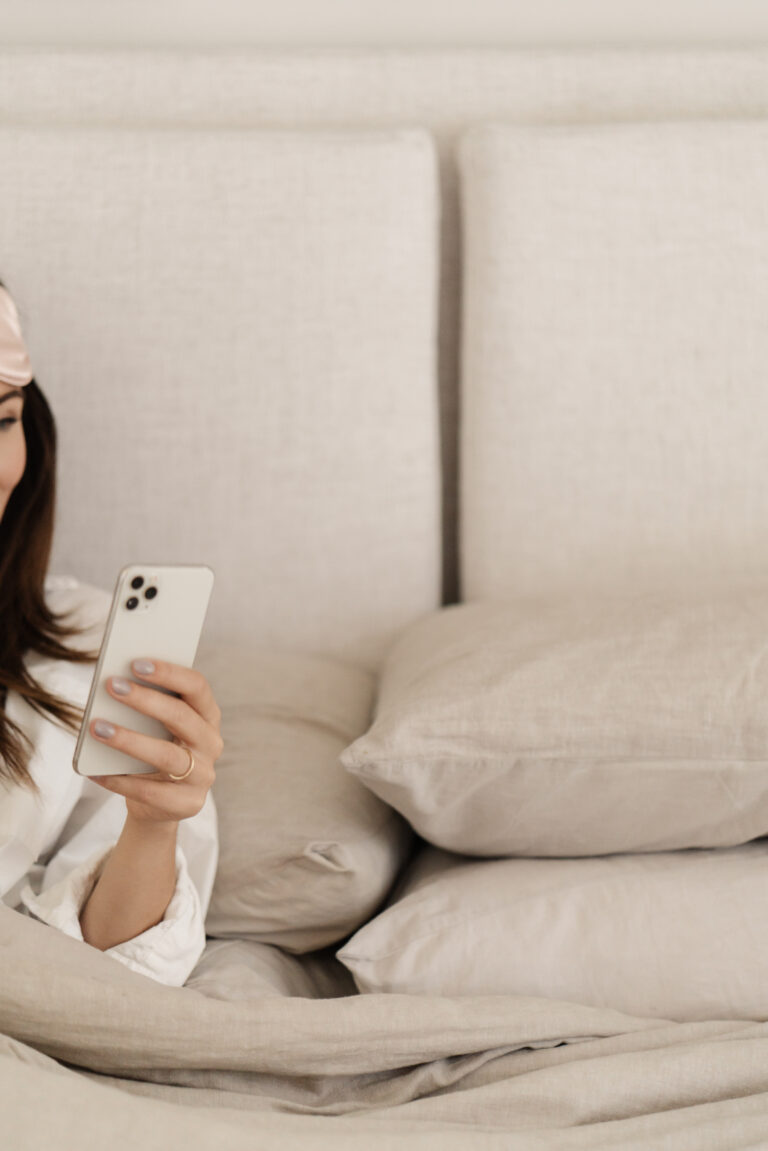 Relaxed woman in bed with a satin eye mask, holding a smartphone, embracing self-care and manifestation practices.