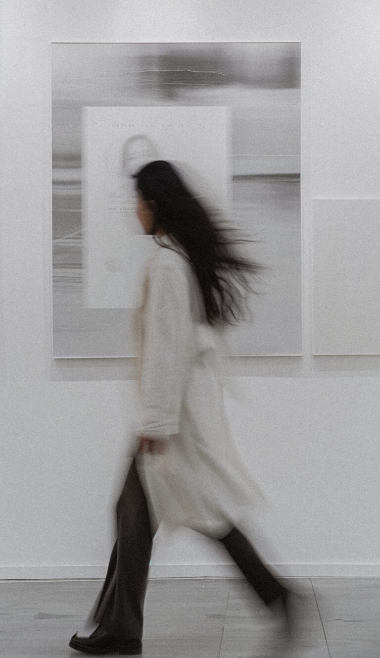 Artistic black and white photo of a woman in motion, wearing white, walking past a gallery wall - embodying main character energy and personal transformation