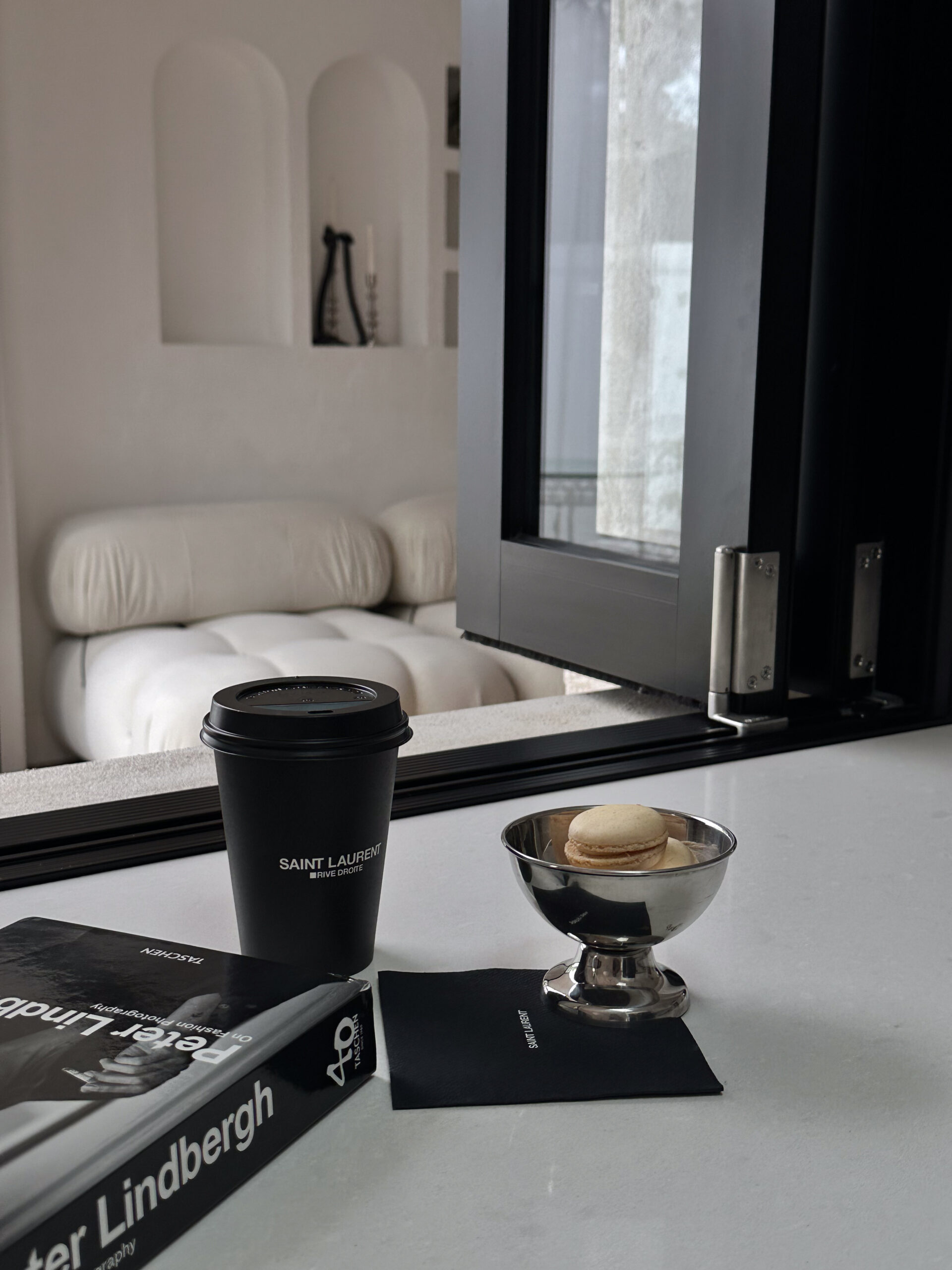 Styled minimalist scene with Saint Laurent coffee cup, fashion book, and macaron on white surface