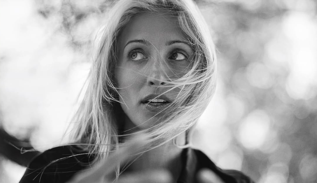 Black and white close-up portrait of Carolyn Bessette Kennedy with windswept blonde hair looking off to the side, wearing a dark top against a bokeh background
